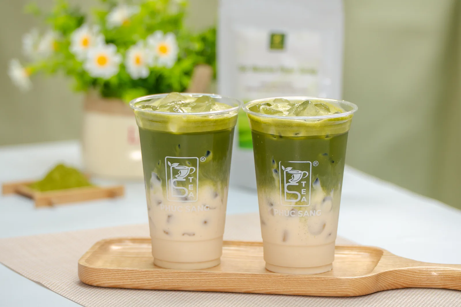 Hai ly matcha latte đá đặt trên khay gỗ, bên cạnh chén bột matcha và phới đánh