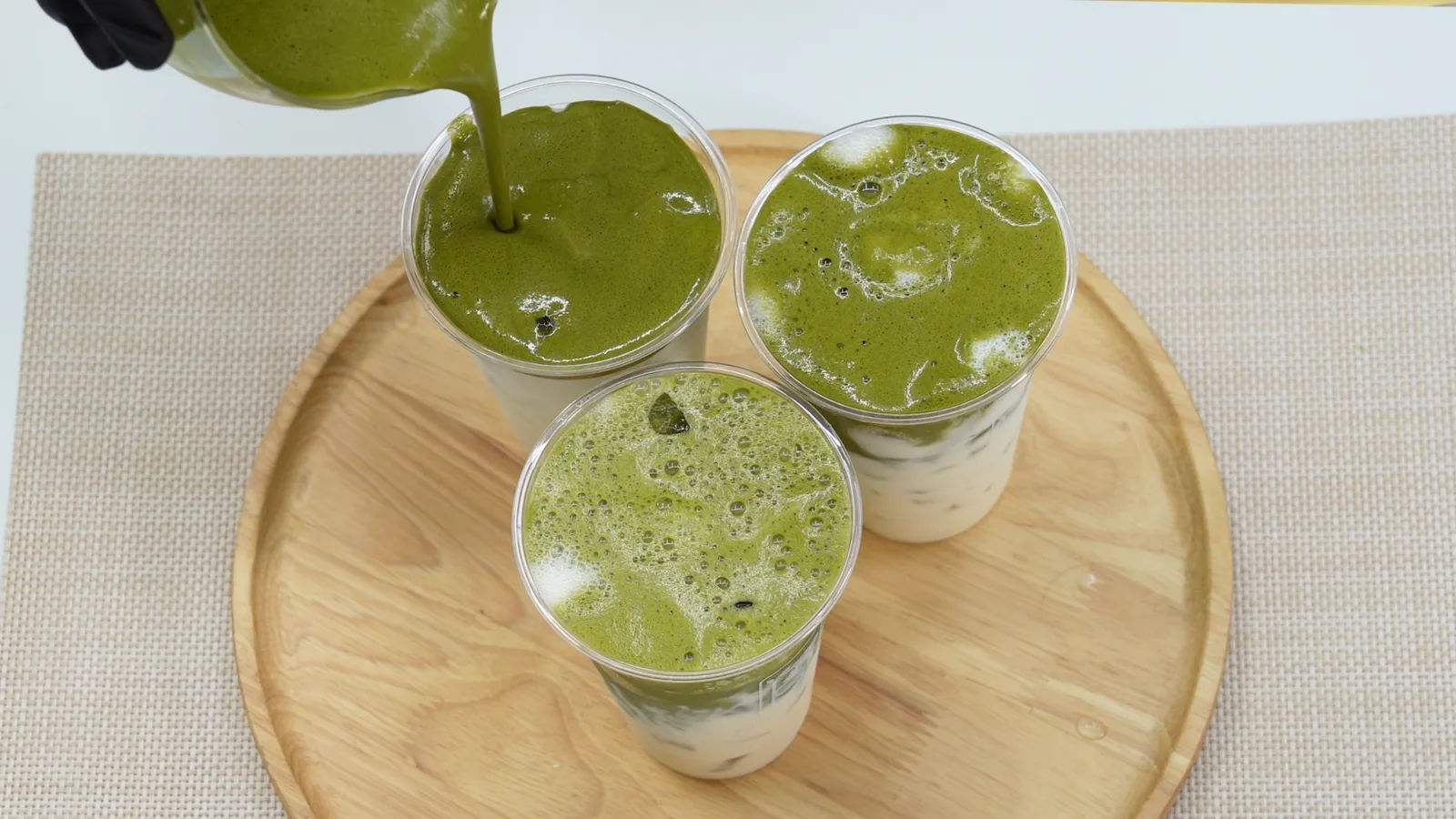 Đang rót matcha đã đánh vào ba ly nhựa đặt trên khay gỗ trong bếp thực hành