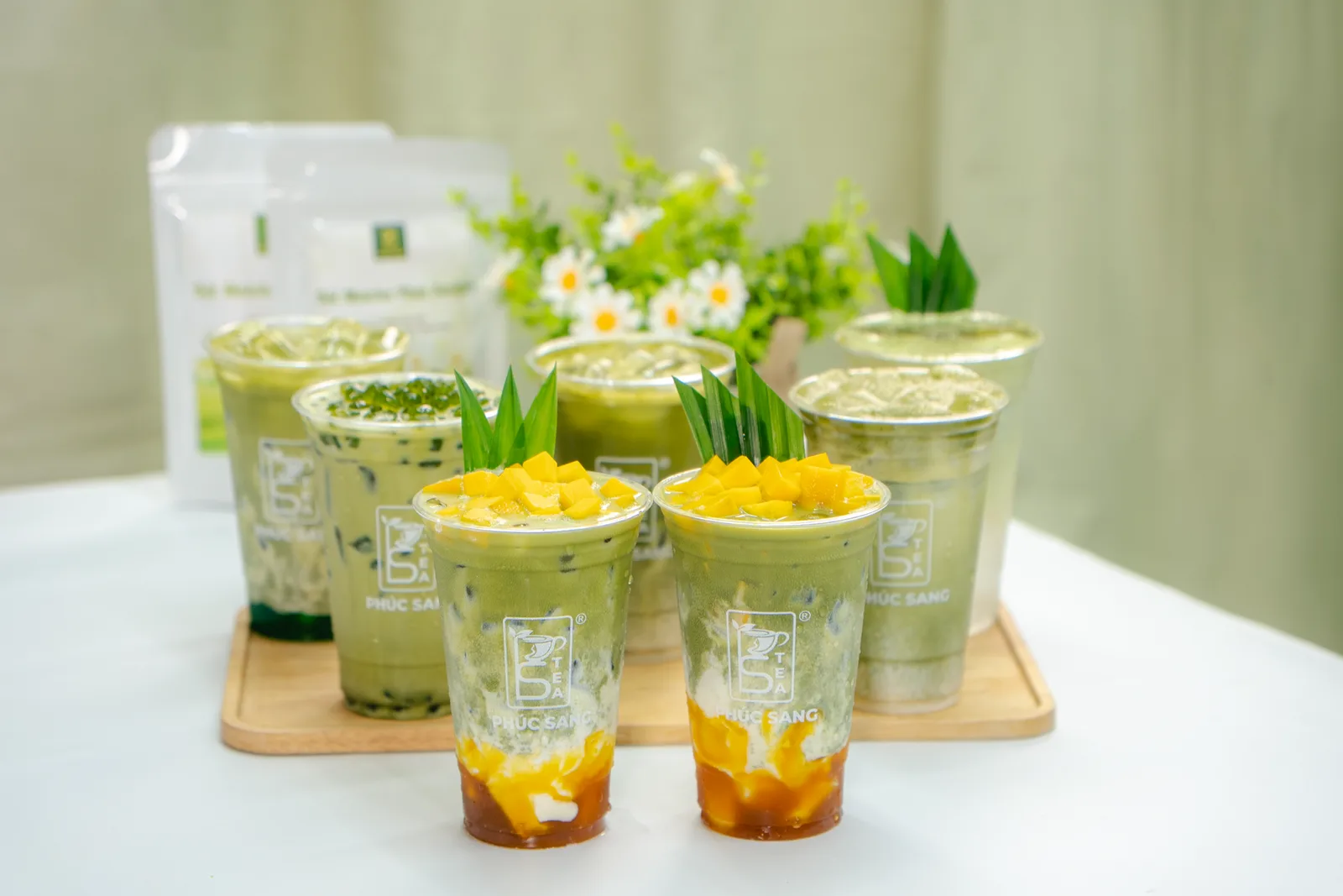 Nhiều ly matcha latte và matcha xoài latte xếp trên khay gỗ cùng bình hoa